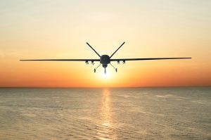 Unmanned military drone patrols the territory at sunset, flying above water surface. The view is straight ahead.
